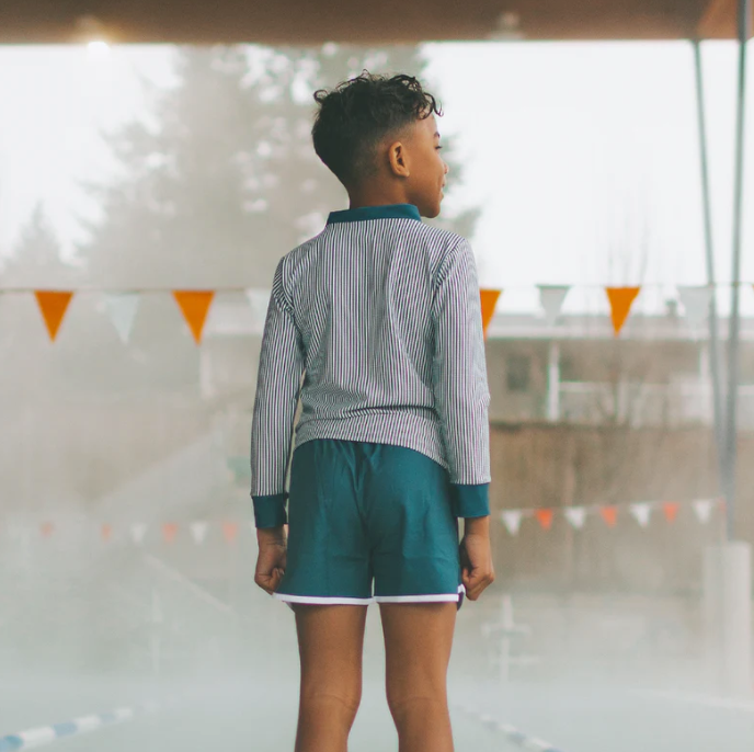 Person wearing a striped shirt and navy swim shorts standing in an indoor pool area.