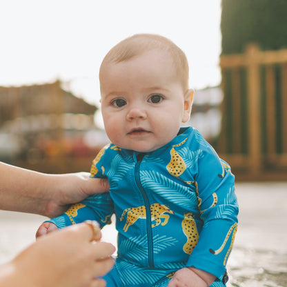 Baby in a blue swimsuit with yellow patterns sitting on a pool ledge, being helped by an adult.