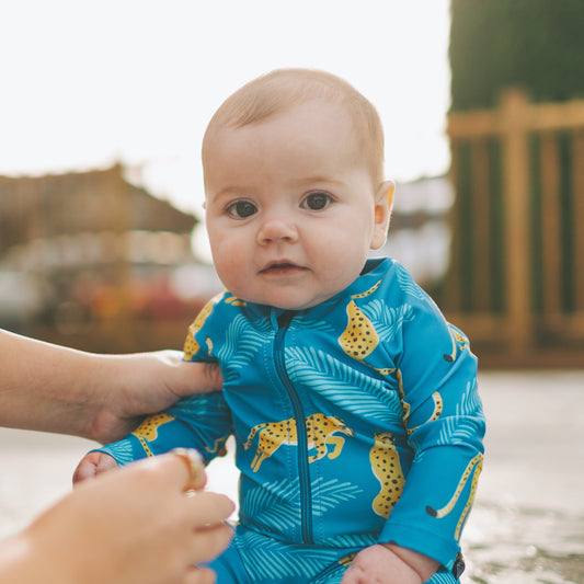 Baby in a blue swimsuit with yellow patterns sitting on a pool ledge, being helped by an adult.