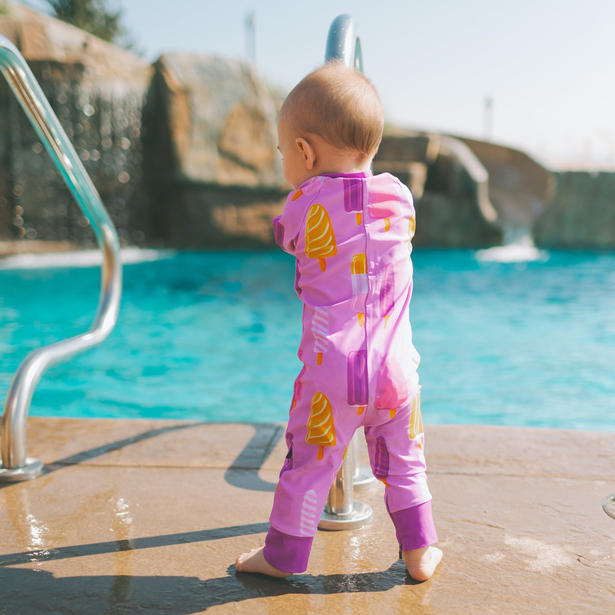 A baby standing on a pool deck wearing a pink one-piece swimsuit with yellow ice cream cone prints, facing a swimming pool with rock formations in the background.