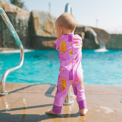A baby standing on a pool deck wearing a pink one-piece swimsuit with yellow ice cream cone prints, facing a swimming pool with rock formations in the background.