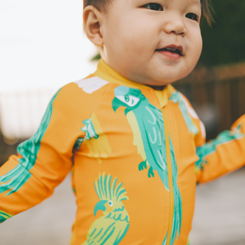 Child wearing an orange onesie with green parrot designs outdoors.