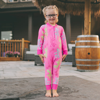 Child in a pink floral onesie standing on a stone patio with a tiki hut in the background.