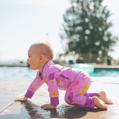 A baby crawling on a wet poolside deck wearing a pink long-sleeve one-piece swimsuit with a colorful abstract pattern.