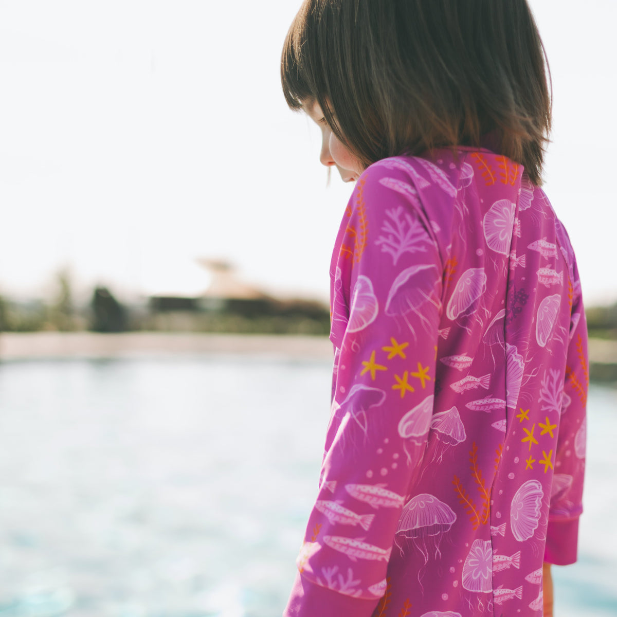 A child wearing a bright pink long-sleeve swimsuit set with a white and orange sea-creature print, standing by a swimming pool.